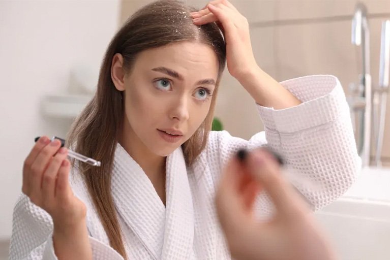 Wanita mengenakan bathrobe putih sedang mengaplikasikan hair serum Kérastase ke kulit kepala untuk perawatan rambut sehat dan berkilau.